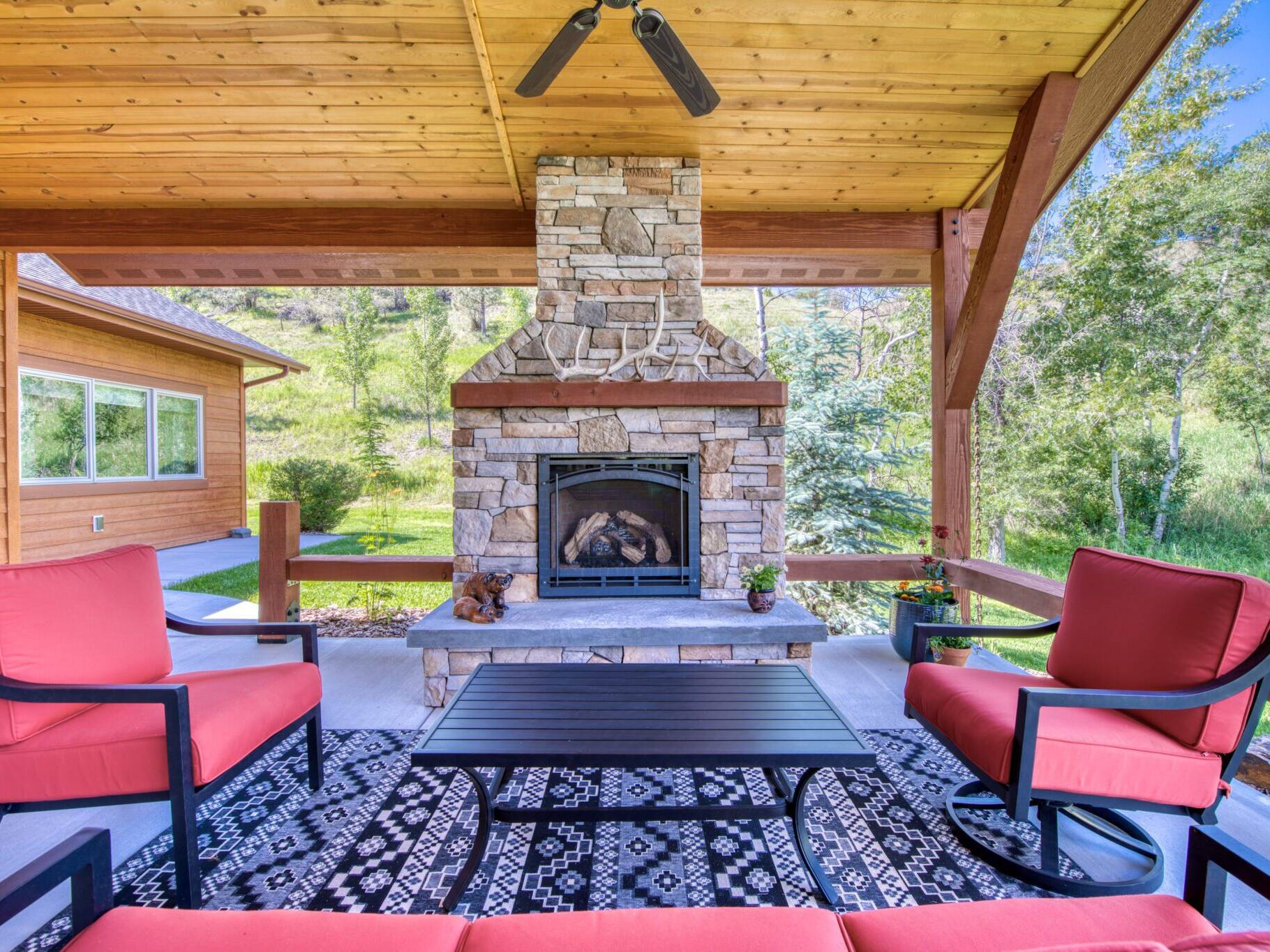 Covered back patio with vaulted wood ceilings, accent beams, a gas fireplace with raised hearth, stone masonry and a mantle in a custom home near Hamilton, MT