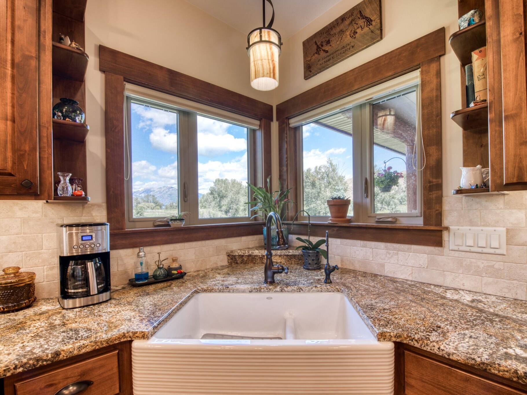 Farm-style kitchen sink in a corner cabinet with granite countertops, tile backsplash and two windows above in a custom home near Hamilton, MT