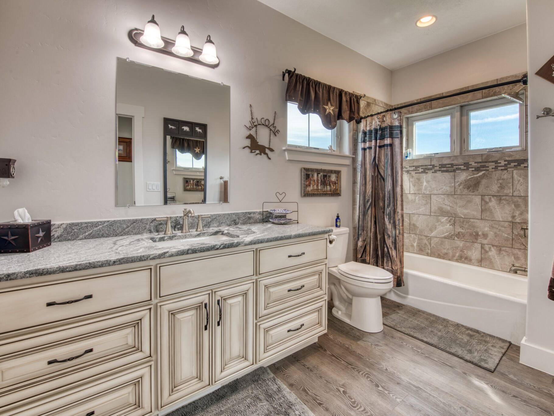 Guest bathroom with LVP flooring, white glazed cabinets, granite countertops, tub/shower with tile surround and windows in a custom home built by Big Sky Builders in Stevensville, MT